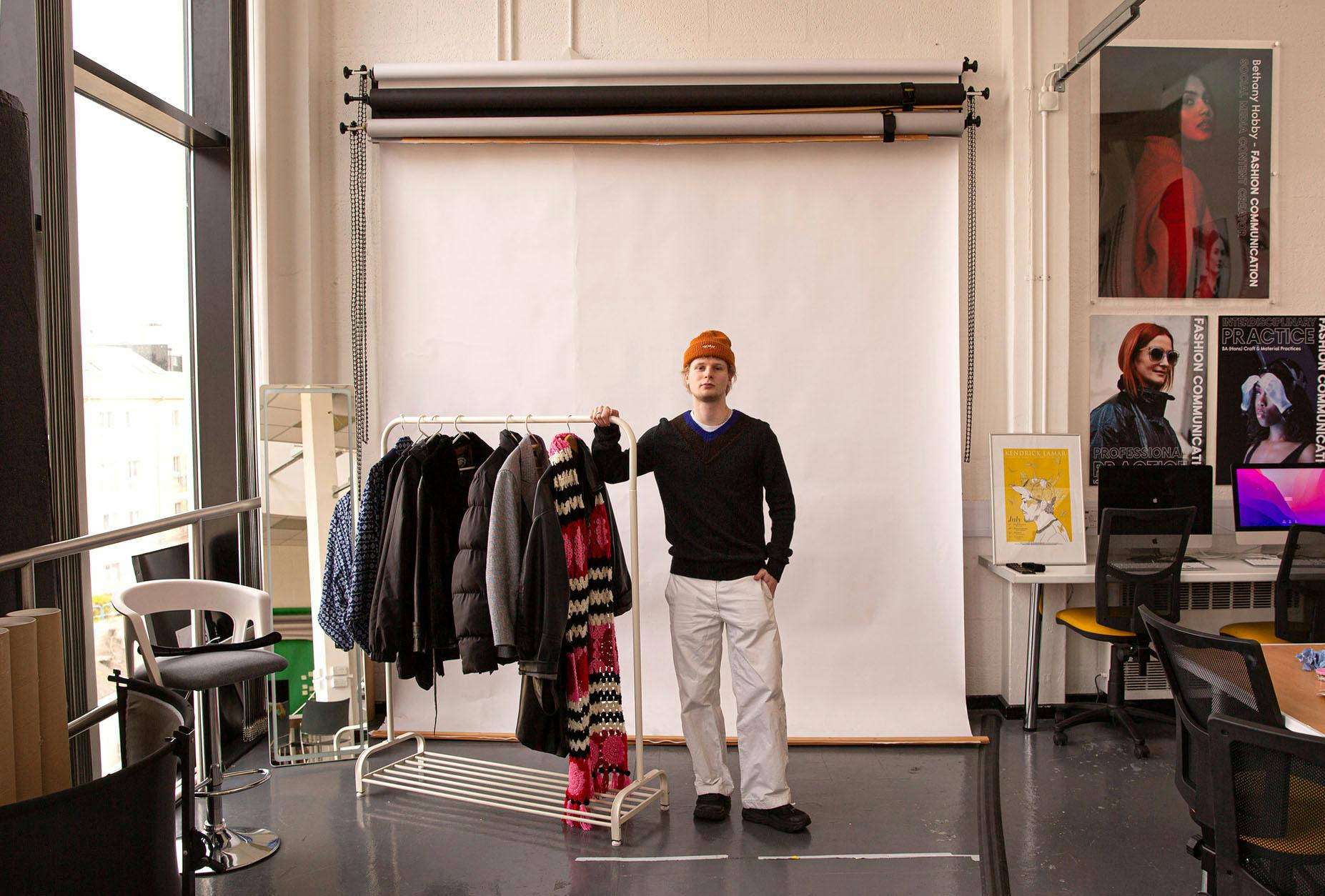 Fashion Communication student Harry Langford stands with a rail of clothes in the studio in front of a white colorama